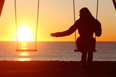 A woman sits on a swing over looking water as the sunsets, with an empty swing beside her.